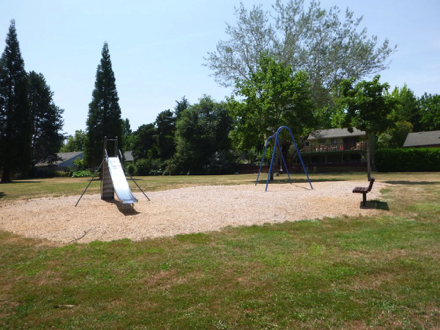 Playgrounds with slide, swings and bench has a bark chip surface – surrounded by grass
